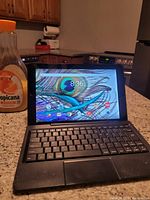Tablet displayed on kitchen countertop with keyboard attached, showing home screen with peacock feather wallpaper.