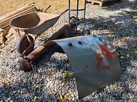 Side view of scrap metal pieces with orange spray paint, rusty pipe, wheelbarrow and dolly on gravel ground