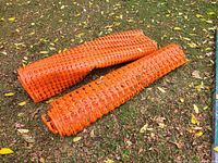 Two orange plastic snow fence rolls lying on grass, showing the rolled mesh with oval holes.