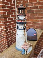 Steel decorative lighthouse figurine with small attached house shown standing next to a baseball cap for size reference against a brick wall background.