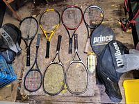 Seven tennis rackets, some grip tape worn or missing, arranged on wooden surface with two racket cases and tennis ball container.