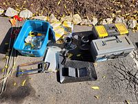 Photo showing blue plastic tub holding various small tools and packaged electrical outlets, alongside metal hardware parts and gray tool box on pavement with leaves around.