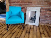 Front view of turquoise leather chair next to vintage white framed hardwood mirror against brick wall.