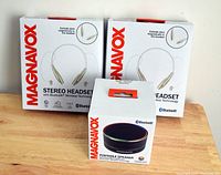 Photo showing two boxed Magnavox stereo Bluetooth wireless headsets and one boxed Magnavox Bluetooth portable speaker on a wooden surface.