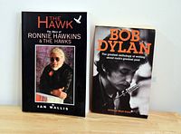 Photo of two softcover books placed upright on a white surface against a plain background. The left book is about Ronnie Hawkins, the right book about Bob Dylan.