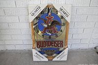 Front view of Budweiser quartz clock in original plastic packaging with visible rips and cardboard corner protectors.