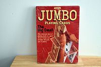 Front view of unopened jumbo deck of playing cards in red box showing the cards arranged as a house of cards design.
