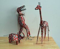 Photo showing three Coca-Cola tin can folk art animal sculptures including elephant, dinosaur, and giraffe on a wooden surface against a white wall.
