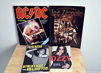 Four music books arranged on a wooden surface: AC/DC Anthology tab book with image of Angus Young playing guitar, Led Zeppelin Drum Techniques tab book with drummer image, Pat Benatar hardcover biography, Ozzy Osbourne softcover biography.