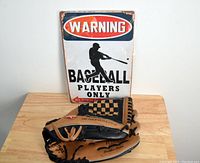 Metal sign with a silhouette of a batter and warning text, placed behind the Rawlings baseball glove on a wooden surface.