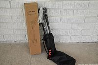 Photo showing TACKLife tripod in original box alongside protective carrying pouch, tripod legs wrapped in plastic.