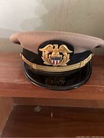 Front view of one US Navy officer hat with beige top, black band, and gold eagle insignia.