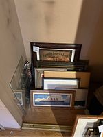 Six pieces of various framed artwork arranged leaning against a wall on a wooden floor, including a vintage travel poster and ship imagery.
