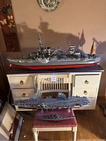 Front and side view of three battleship replicas displayed on and in front of a cream colored desk and chair. The largest replica is about 80 inches long on top of the desk, with the aircraft carrier and smaller battleship below on the chair.