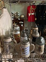 Overview of ten beer steins arranged together on a wire shelf with some historical maritime clothing and hats in background.