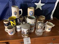 Top-down view of entire collection of 16 assorted beer steins and mugs on wooden surface with flag backdrop