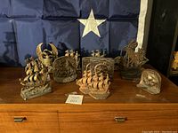 Display of all 13 miscellaneous heavy military-themed bookends arranged on wooden surface in front of a blue flag with white star. Several bookends depict ships and eagle designs.