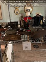 Wide view showing multiple items on metal shelving including lighthouse lamp, ship propeller, relief plaque, firefighter and shield emblem sculptures, military hats, and jackets.