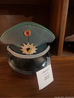 Front view of East German military hat showing green piping, red cockade, silver emblem with shield, and black visor with silver braid.