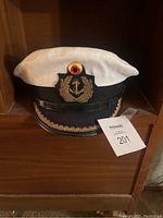 White and black West German Navy officer hat with anchor insignia and gold embroidery on the visor, placed on a wooden shelf with auction tag