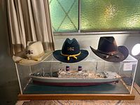 Three western hats on display case with model ship inside, from left: light tan, dark blue, dark brown.