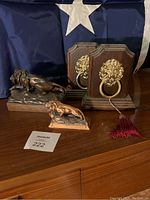 Photo showing two lion sculptures and a pair of lion head door knockers with tassel, placed on wooden surface with blue fabric backdrop