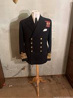 Full front view of WWII U.S. Navy dark navy dress jacket on wooden stand with shirt and tie underneath, showing gold buttons and ribbon bars.