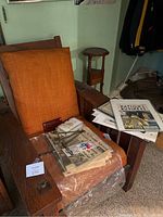 Photo of orange upholstered wooden armchair with vintage newspapers, magazines, and items on and around it.