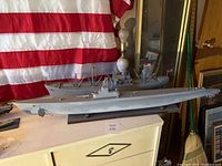 Two gray replica navy ships on top of a beige dresser with an American flag backdrop. The first is a submarine and the second is a cargo/supply ship with masts and flags.