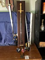Photo showing three swords with wooden plaque in center, metal miniature flags, and figurine in front.