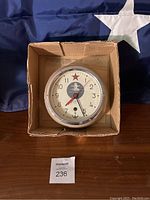 Clock inside cardboard box on wooden table showing front view with red star, lighthouse submarine emblem, and anchor symbol.