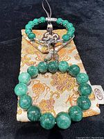 Two green dyed jade bracelets displayed on silk brocade pouches, showcasing bead size difference, silver tone charms, and threaded cord details.