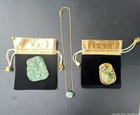 Two carved natural stone pendants each resting on black pouches with gold drawstrings, and a gold tone necklace with light green stone pendant laid between them.