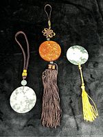 Photo showing all three medallions laid on a black background: two natural stone medallions with cords and tassels, and one carved wooden medallion with brown tassel.