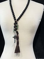 Full view of the black obsidian bead necklace worn on a mannequin torso, showing the overall length, bead size, and arrangement of pendants including the tassel and cross charm.