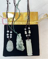 Three jade pendant necklaces on black and gold pouch, showing front and side views of pendants and cords
