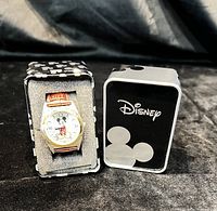 Mickey Mouse wristwatch inside a foam insert in a black and white metal box with Disney branding and Mickey Mouse silhouette pattern on lid.