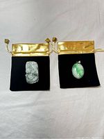 Both pendants on black velvet pouches with gold drawstrings, showing the scale and color contrast