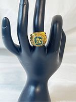 Front view of the gold tone Oakland Athletics championship ring displayed on a black hand-shaped stand showing size and design.