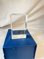 Front view of clear rectangular prism glass paperweight on blue box, showing height and clarity.