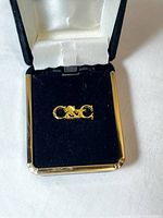 Gold tone C&C initial pin resting in black velvet, satin-lined jewelry box with gold trim, showing front view of pin.