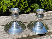 Pair of antique sterling silver squat candlesticks displayed outdoors on a wooden surface showing their symmetrical shape and reflective silver finish.