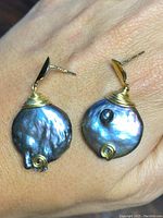 Pair of large black pearl earrings with gold wire wrappings on a hand background under bright lighting, highlighting the pearls' iridescence and gold details.