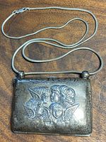 View of the sterling silver rectangular compact or smoking case suspended from a silver snake chain necklace, showing the embossed angelic faces on the front with detailed hair and wings.