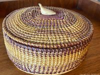 Full view of the closed round woven basket with carved bird finial on top, showing weaving pattern and colors.