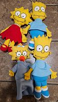 Four plush dolls from The Simpsons, including Lisa, Bart, Maggie, and Milhouse plush toys.