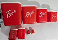 Red mid-century lustre ware canisters labeled Flour, Sugar, Coffee, Tea, Salt, Pepper, plus matching salt and pepper shakers arranged on a white surface.