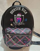 Front view of black Monster High Alumni mini backpack featuring skull logo with pink sequin M and 2010 Alumni text, plus front zippered pocket with plaid pattern and chain detail.