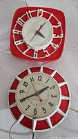 Front view of both mid century wall clocks; one square red clock with white wire frame, one round red and white clock with black numerals and spokes.