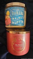 Photo of two scented candles: Banana Walnut Muffin and The Perfect Christmas with visible labels and gold lids.